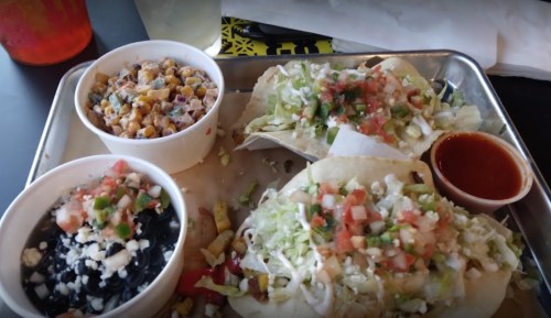A tray with three tacos topped with lettuce and tomatoes, a bowl of corn salad, and a small cup of sauce.