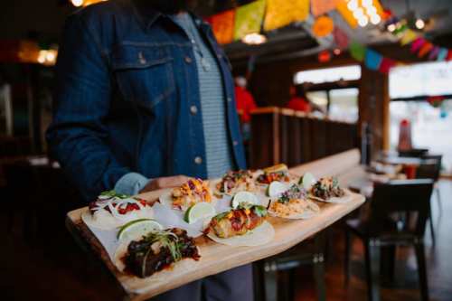 A person holds a wooden platter with various colorful tacos, garnished with lime, in a festive restaurant setting.