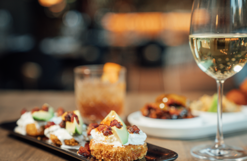 A plate of appetizers with creamy toppings, garnished with herbs, alongside a glass of white wine and a cocktail.