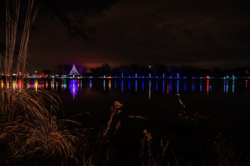 A serene night scene of a lake reflecting colorful holiday lights and a decorated tree in the distance.