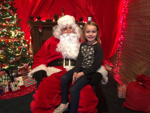 A young girl sits on Santa's lap in a festive room decorated with a Christmas tree and holiday lights.