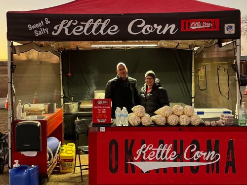 A kettle corn stand with two vendors smiling behind a table filled with popcorn bags and drinks.