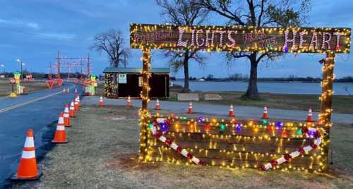 A festive display with lights and decorations near a lake, featuring a sign that reads "Lights from the Heart."