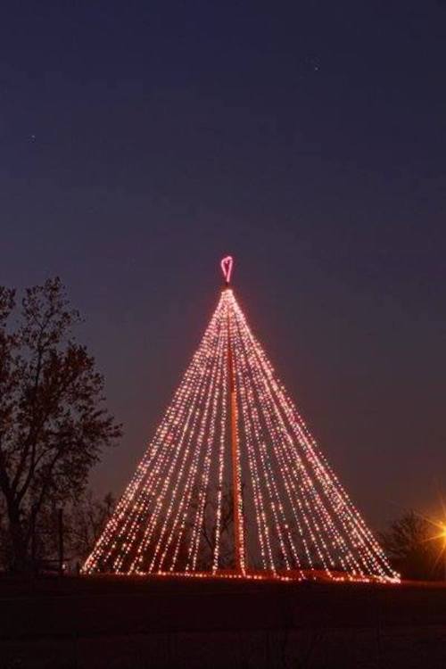 A tall Christmas tree made of lights, glowing against a twilight sky, with a heart-shaped topper.