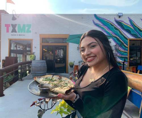 A smiling young woman holds a large quesadilla on a plate outside a colorful restaurant.