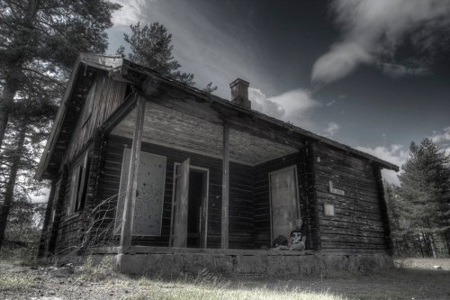 A weathered wooden cabin in a desolate landscape, with an overcast sky and sparse trees surrounding it.