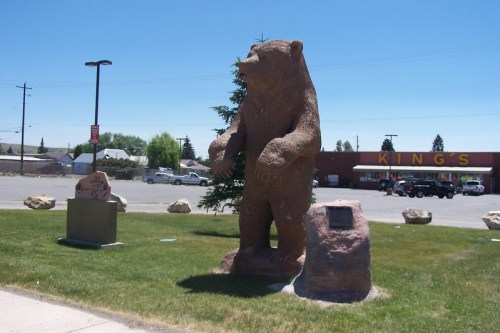 A large bear statue stands on grass, with a plaque nearby and a store in the background under a clear blue sky.