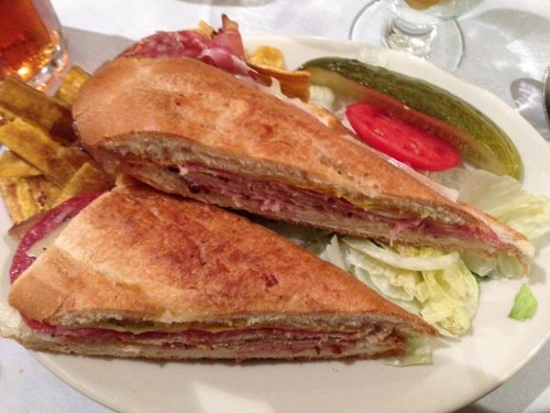 A sandwich cut in half, filled with meats, cheese, and mustard, served with pickles, tomato, and lettuce on the side.
