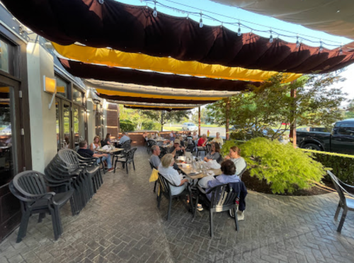 Outdoor dining area with patrons seated at tables under colorful canopies, surrounded by greenery and a parking lot.
