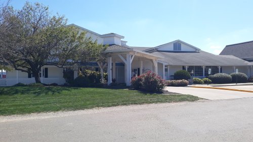 A sunny view of a building with a porch, surrounded by greenery and flower beds.
