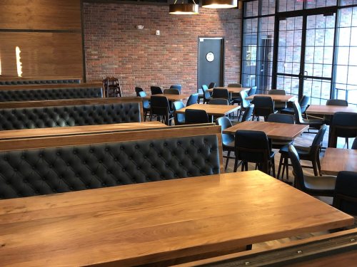 Interior of a restaurant featuring wooden tables, blue chairs, and a brick wall, with large windows letting in light.