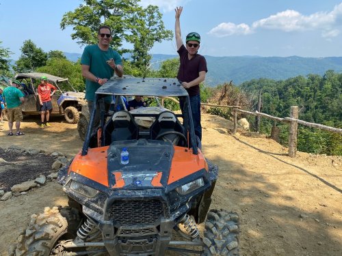Two men stand on an ATV in a scenic outdoor setting, with mountains in the background and a clear blue sky.