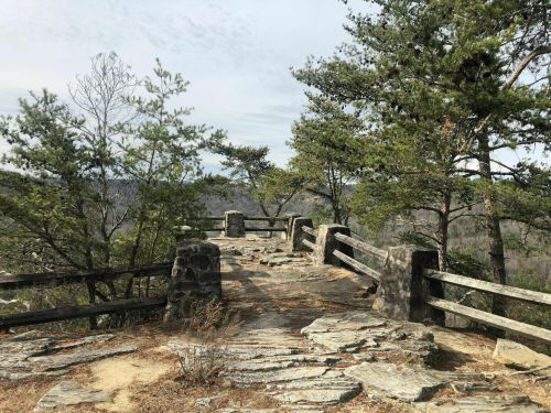 A rocky overlook surrounded by trees, featuring a stone railing and a scenic view of distant hills.
