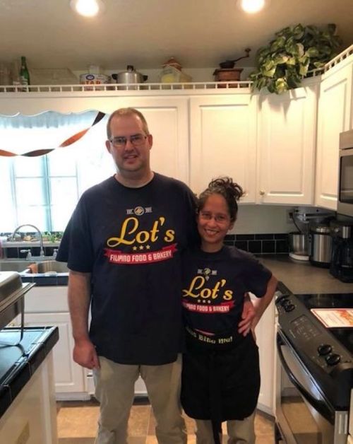 A man and a woman stand in a kitchen, both wearing matching t-shirts for "Lot's Filipino Food & Bakery."