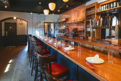 A spacious restaurant bar with wooden seating, a long counter, and shelves stocked with drinks against a brick wall.