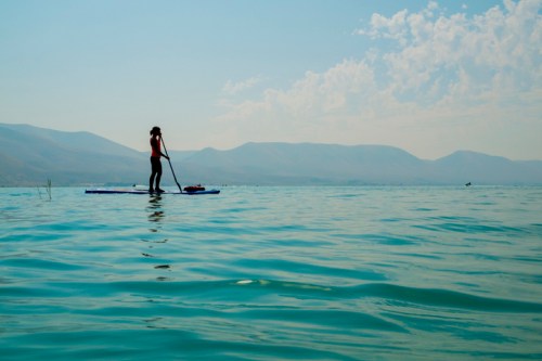 A person paddleboarding on calm water with mountains in the background under a clear blue sky.