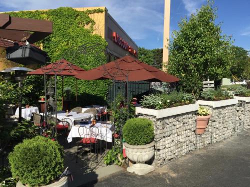 Outdoor dining area with tables under umbrellas, surrounded by greenery and stone walls, next to a restaurant.