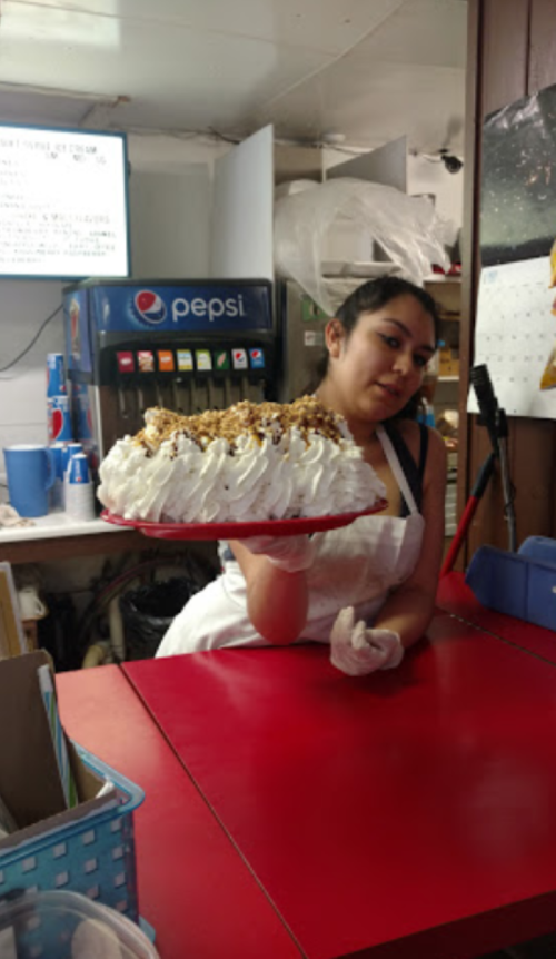 A woman in an apron holds a large, decorated cake in a kitchen setting with a soda machine in the background.