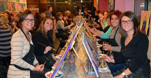 A group of women smiling and painting at easels in a lively art studio setting.
