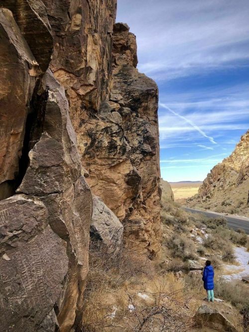 A person in a blue coat stands near large rock formations beside a road, with a vast landscape and sky in the background.