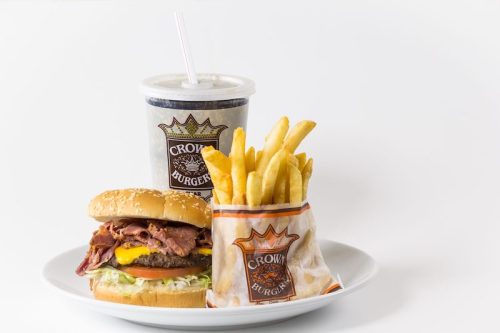 A plate with a burger topped with bacon, a side of fries in a bag, and a drink with a straw.