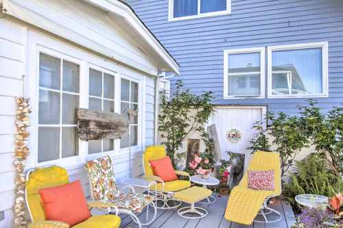 A cozy outdoor seating area with yellow chairs and colorful cushions, surrounded by greenery and a light blue house.