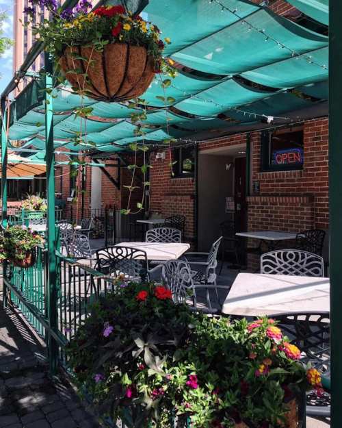 Outdoor dining area with tables, hanging flower baskets, and a bright "OPEN" sign on a sunny day.