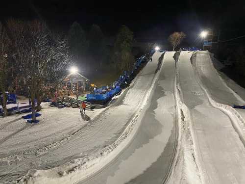 A snowy hill with multiple sledding lanes, illuminated by lights, and a cozy lodge in the background at night.