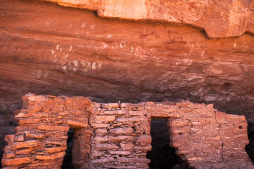 Ancient stone ruins beneath a rocky overhang, featuring faded petroglyphs on the cliff above.