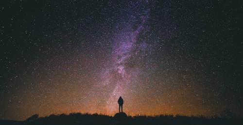A silhouette of a person stands on a hill, gazing up at a starry night sky filled with the Milky Way.