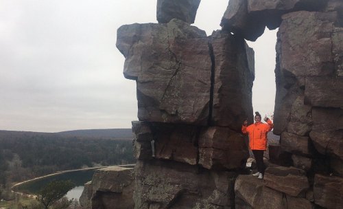 A person in an orange jacket stands between two large rock formations, overlooking a scenic landscape with a lake.