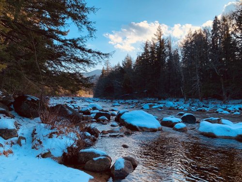 A serene winter landscape featuring a river surrounded by snow-covered rocks and trees under a blue sky.