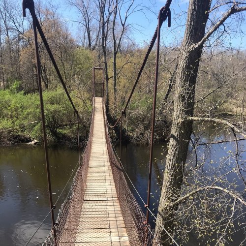 A wooden suspension bridge spans a calm river, surrounded by lush greenery and trees under a clear blue sky.