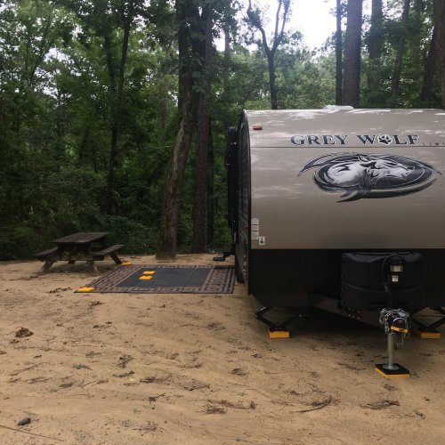 A Grey Wolf travel trailer parked in a wooded area, with a picnic table nearby and trees in the background.