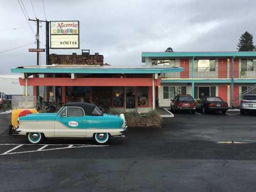 A vintage turquoise and white car parked in front of the Atomic Motel, featuring a retro sign and colorful building.