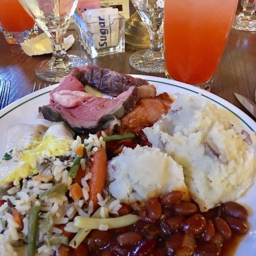 A plate of food featuring sliced meats, mashed potatoes, rice with vegetables, and baked beans, with drinks in the background.
