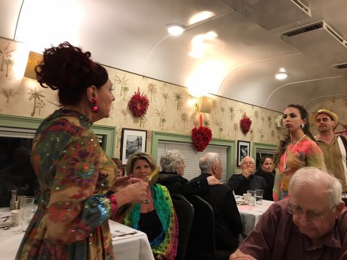 A lively restaurant scene with diners, two performers in colorful outfits, and a woman speaking animatedly.