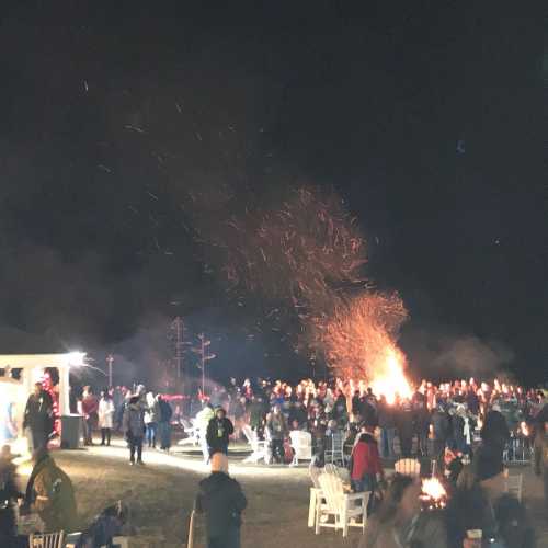 A large crowd gathers around a bonfire at night, with sparks flying into the dark sky.