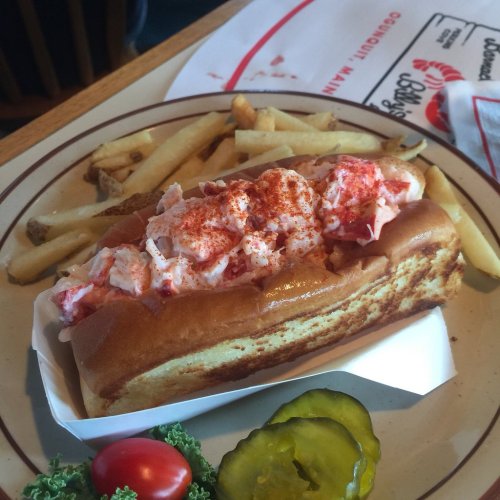 A lobster roll topped with lobster salad, served with fries and garnished with pickles and cherry tomatoes on a plate.