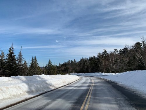 A winding road through a snowy landscape, flanked by snowbanks and evergreen trees under a clear blue sky.
