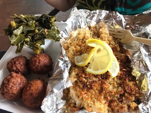 A plate featuring baked fish with lemon, collard greens, and two golden cornbread muffins.