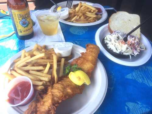 A plate with fried fish, fries, coleslaw, and a slice of bread, alongside a drink and another plate in the background.