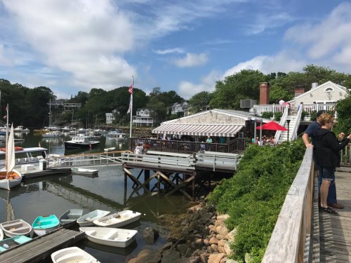 A scenic waterfront view featuring boats, a restaurant with outdoor seating, and people enjoying the area.