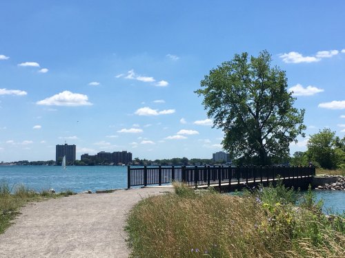 A scenic view of a wooden pier extending into a blue lake, surrounded by greenery and buildings under a clear sky.