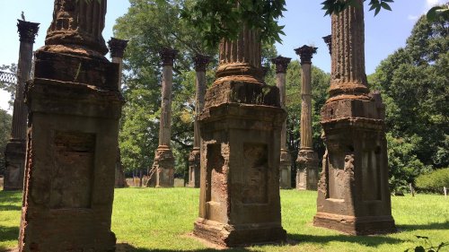 Ruins of tall, weathered columns surrounded by lush greenery under a clear blue sky.