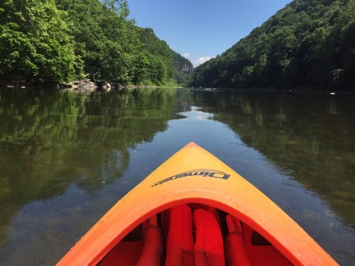 A kayak's bow on calm water, surrounded by lush green hills under a clear blue sky.