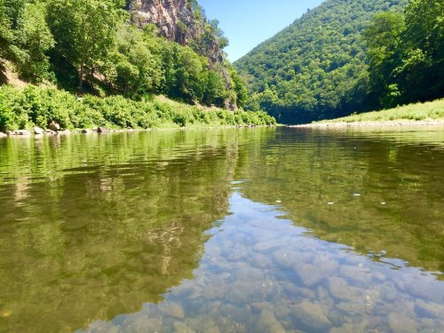 A serene river scene with clear water reflecting lush green hills and trees on either side.
