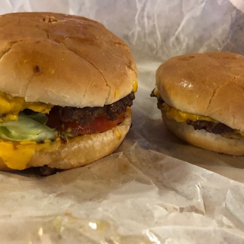 Two cheeseburgers on a paper wrapper, one larger with lettuce and tomato, the other smaller and simpler.