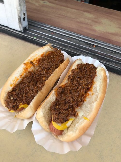 Two hot dogs in buns topped with a thick, dark chili sauce and mustard, served on a paper tray.