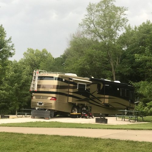 A large, luxurious RV parked in a green, wooded area with picnic tables nearby.